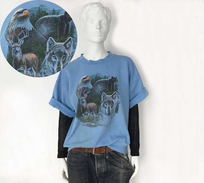 May include: A light blue t-shirt with a wildlife graphic depicting an eagle, bear, deer, and wolf. The sleeves are rolled up, and it's worn over a black long-sleeved top. The mannequin is wearing blue jeans and a brown belt.