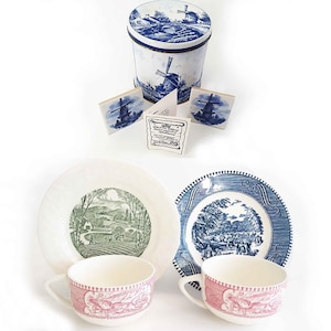 May include: Two teacups and saucers featuring a blue and white windmill design. The teacups have a pink and white striped band around the top. The saucers have a blue and white design with a windmill and a landscape. There is also a blue and white tin with a windmill design and two blue and white coasters with a windmill design.