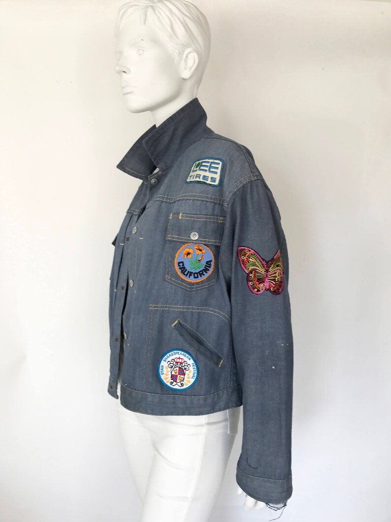 K&ouml;nnte beinhalten: Eine blaue Jeansjacke mit verschiedenen Patches, darunter ein Schmetterling, eine Blume und ein Logo mit der Aufschrift "Lee Tires".