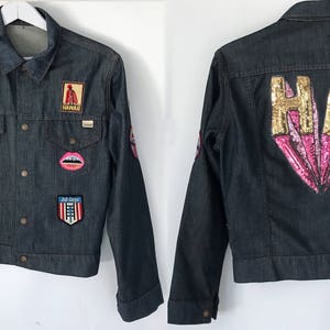 Peut inclure: Une veste en jean bleu fonc&eacute; avec divers patchs et embellissements. Le dos de la veste pr&eacute;sente un grand c&oelig;ur en sequins rose et or avec les lettres "HA" en sequins dor&eacute;s.