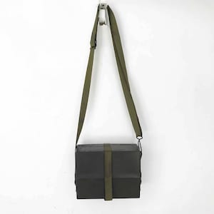Peut inclure: Une boîte rectangulaire noire avec une sangle en toile verte. La boîte a un rabat qui se ferme avec un fermoir en métal. La sangle est réglable et possède un crochet en métal.