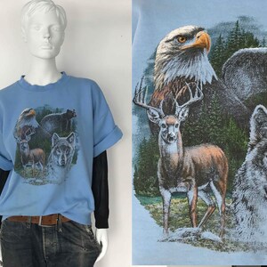 May include: Pale blue graphic t-shirt featuring an eagle, bear, deer, and wolf. The shirt has rolled-up sleeves and is worn over a black long-sleeved shirt. The graphic depicts the animals in a forest setting.