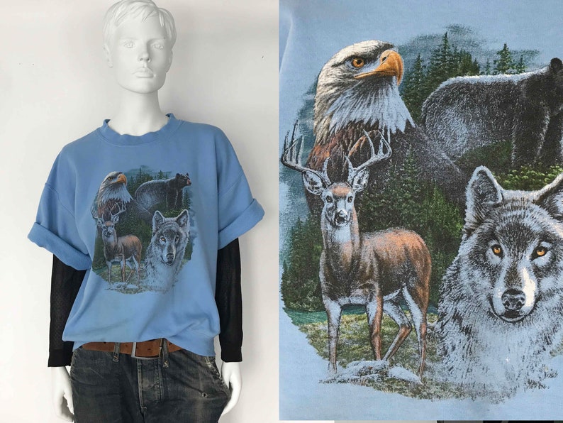 May include: Pale blue graphic t-shirt featuring an eagle, bear, deer, and wolf. The shirt has rolled-up sleeves and is worn over a black long-sleeved shirt. The graphic depicts the animals in a forest setting.
