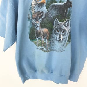 May include: Pale blue short-sleeved sweatshirt with a wildlife design. The graphic depicts an eagle, a deer, a wolf, and a bear. The animals are set against a backdrop of trees and foliage.