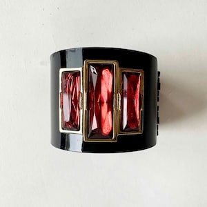 May include: A wide black cuff bracelet featuring three rectangular, faceted red stones set in gold-toned metal frames. The central stone is larger than the two flanking stones, and the entire piece has a smooth, glossy finish on the black band.