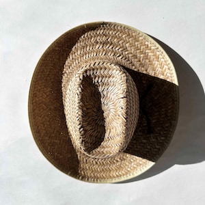 Puede incluir: Sombrero de paja tejido de color beige natural, con ala ancha y copa estructurada. El sombrero tiene una textura tejida. El sombrero está sobre una superficie blanca, con una sombra proyectada sobre el ala. El diámetro del ala es de unos 30 cm.