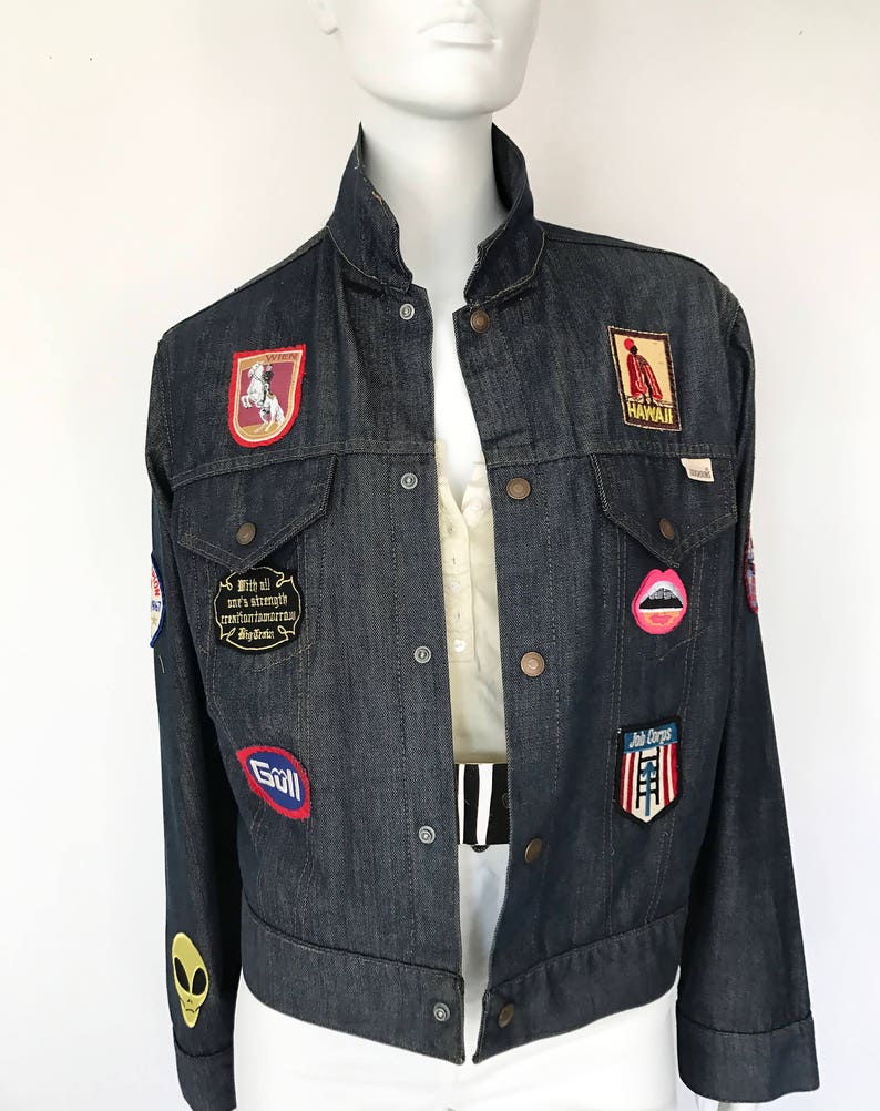 Peut inclure: Une veste en jean bleu fonc&eacute; avec une vari&eacute;t&eacute; de patchs, dont un patch rouge, blanc et bleu avec le texte "With all our strength and courage we confront tomorrow My Dreams", un patch rouge, blanc et bleu avec le texte "Join Corps", un patch rouge, blanc et bleu avec le texte "Goll", un patch rouge, blanc et bleu avec le texte "Hawaii", un patch rouge, blanc et bleu avec un cheval et un cavalier, et un patch rose avec une paire de l&egrave;vres.