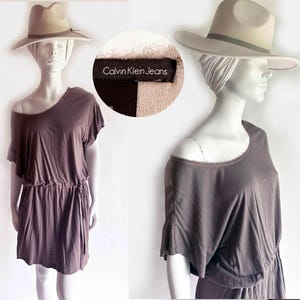 May include: A dark mauve short-sleeved dress with a wide neckline and a drawstring waist. The garment features a Calvin Klein Jeans label. The outfit is completed by a beige fedora hat with a dark band, displayed on a white mannequin.