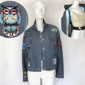 Peut inclure: Une veste en jean bleue avec divers patchs colorés, dont un hibou, un triangle et un globe. La veste a une fermeture éclair et deux poches avant.