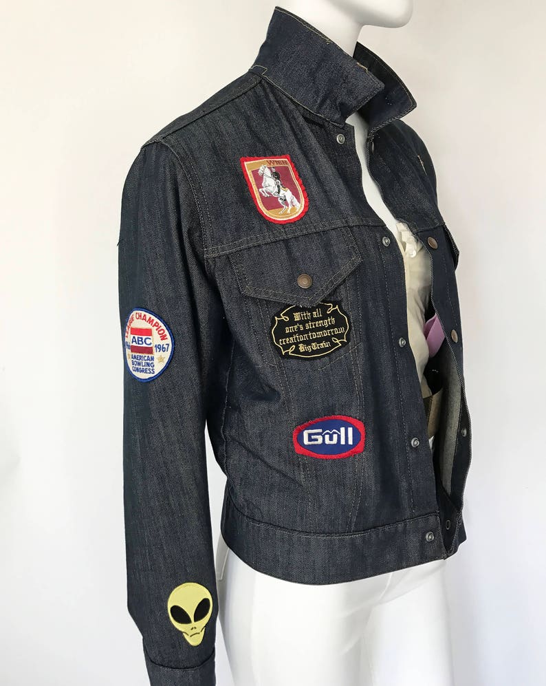 Peut inclure: Une veste en jean bleu fonc&eacute; avec divers patchs, dont un cheval et un cavalier, un ovale rouge et bleu avec le mot "Goll" et un patch blanc avec le texte "With all our strength creation tomorrow Big Dreams". La veste a &eacute;galement un patch d'alien vert sur la manche gauche et un patch rouge, blanc et bleu avec le texte "ABC 1967 American Bowling Congress".
