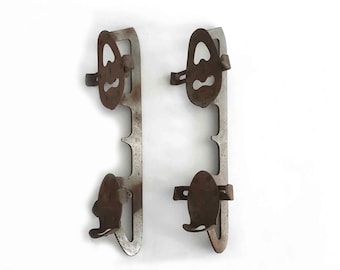 May include: A pair of vintage ice skate blades. Each blade has a metal frame with two oval, rusty brown holders. The blades are attached to the frame with rivets. The frames are a dull silver colour and have a series of cutouts.