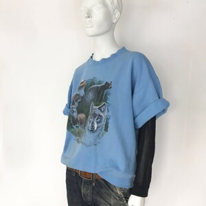 May include: Pale blue sweatshirt with a wildlife print depicting an eagle, bear, deer, and wolf. The sleeves are rolled up, revealing a black long-sleeved top underneath. The mannequin also wears blue jeans and a brown belt.