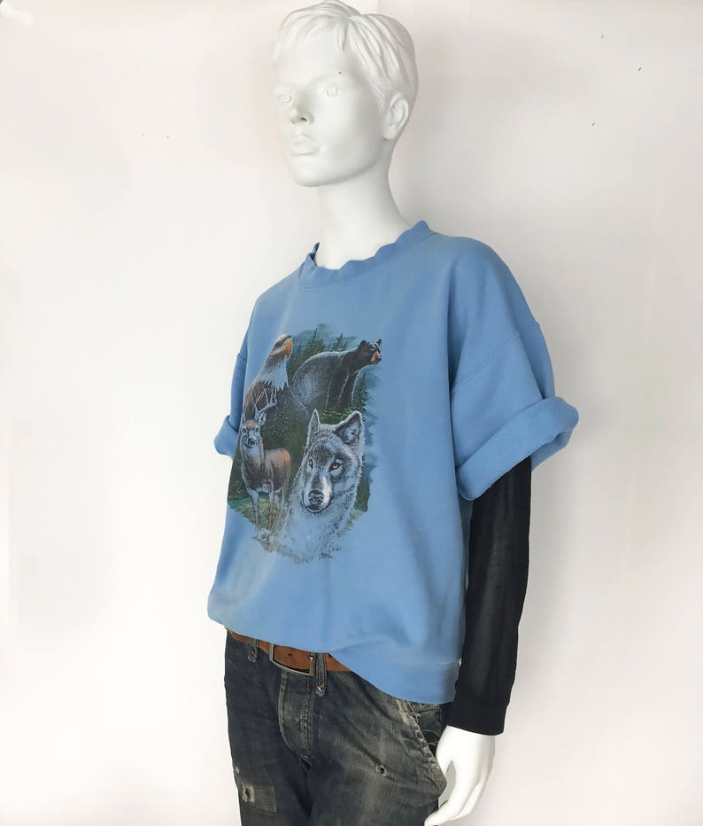 May include: Pale blue sweatshirt with a wildlife print depicting an eagle, bear, deer, and wolf. The sleeves are rolled up, revealing a black long-sleeved top underneath. The mannequin also wears blue jeans and a brown belt.