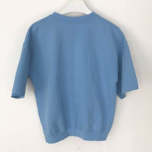 May include: A pale blue short-sleeved sweatshirt hanging on a white hanger. The sweatshirt features a crew neck and a loose fit. The sleeves are short and wide. The material looks to be a soft, medium-weight fabric.