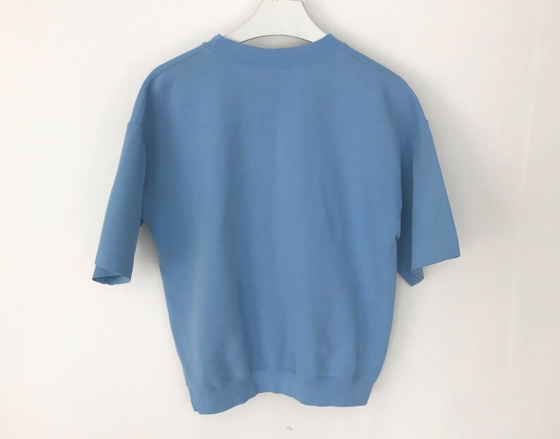 May include: A pale blue short-sleeved sweatshirt hanging on a white hanger. The sweatshirt features a crew neck and a loose fit. The sleeves are short and wide. The material looks to be a soft, medium-weight fabric.