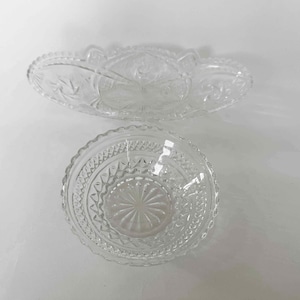 May include: A clear glass oval dish with a scalloped edge and a starburst pattern, paired with a round clear glass bowl featuring a textured diamond and sunburst design. Both pieces exhibit intricate pressed glass detailing and reflective surfaces.