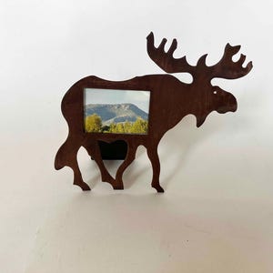 May include: A rustic metal moose-shaped picture frame. The frame is a dark brown color and holds a landscape photo. The moose has large antlers and is mounted on a white wall. The frame is designed to stand on a flat surface.
