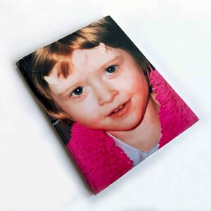 May include: A rectangular photo print featuring a young child with fair skin, blue eyes, and light brown hair. The child is wearing a pink fuzzy garment. The background is white.