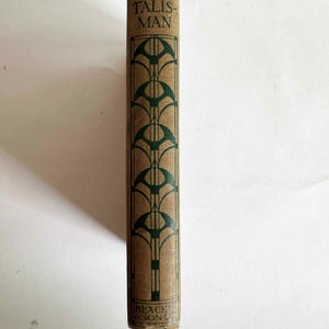 May include: A vintage book spine with the title "THE TALISMAN" in green lettering. The cover is tan with a repeating green art deco design. The publisher's name is visible at the bottom.