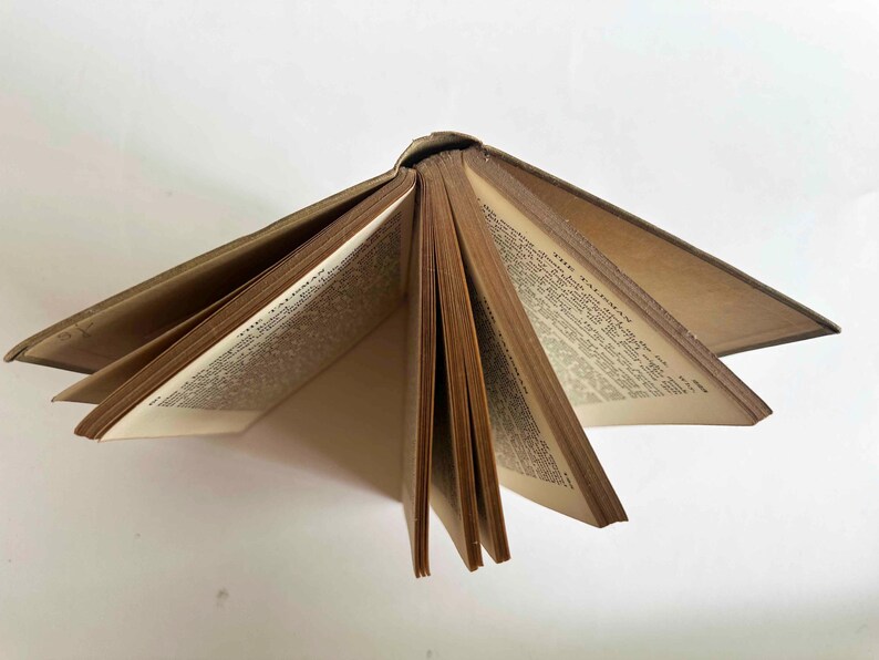 May include: An open antique book with aged, yellowed pages. The book's spine is visible, and the pages are fanned out, displaying printed text. The cover appears to be a worn, light brown color. The book is set against a white background.