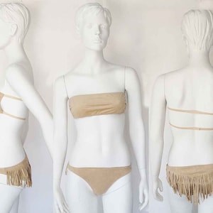 May include: Tan suede bikini set. The top is a bandeau style, and the bottoms are a classic cut with fringe detailing. The bikini is displayed on a mannequin, showcasing the design from multiple angles.