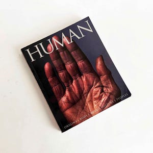 May include: A hardcover book titled "HUMAN" with a close-up image of a hand on the cover. The hand is a reddish-brown color, and the background is dark blue. The book's subtitle reads "ORIGINS | BODY | MIND | CULTURE | PEOPLES."