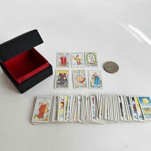 Puede incluir: Una baraja de cartas de tarot en miniatura con una caja negra con interior rojo. Se muestran varias cartas, con una moneda para comparar el tamaño. Las cartas presentan ilustraciones coloridas y están dispuestas sobre una superficie blanca.