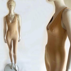 May include: A tan-colored jumpsuit with a plunging V-neckline and fitted silhouette. The one-piece garment features thin straps and a form-fitting design, displayed on a mannequin. The jumpsuit is made of a smooth, possibly stretchy material.