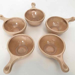 May include: Five beige ceramic soup bowls with handles. The bowls are round with a speckled finish and a small handle on one side. The bowls are arranged in a circular pattern against a white background.