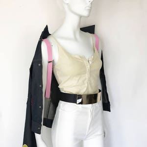 Peut inclure: Un mannequin portant un d&eacute;bardeur blanc, un pantalon blanc, une ceinture noire avec une boucle dor&eacute;e et une veste en jean bleu fonc&eacute; avec des bretelles roses. La veste a un motif extraterrestre sur le c&ocirc;t&eacute; gauche.