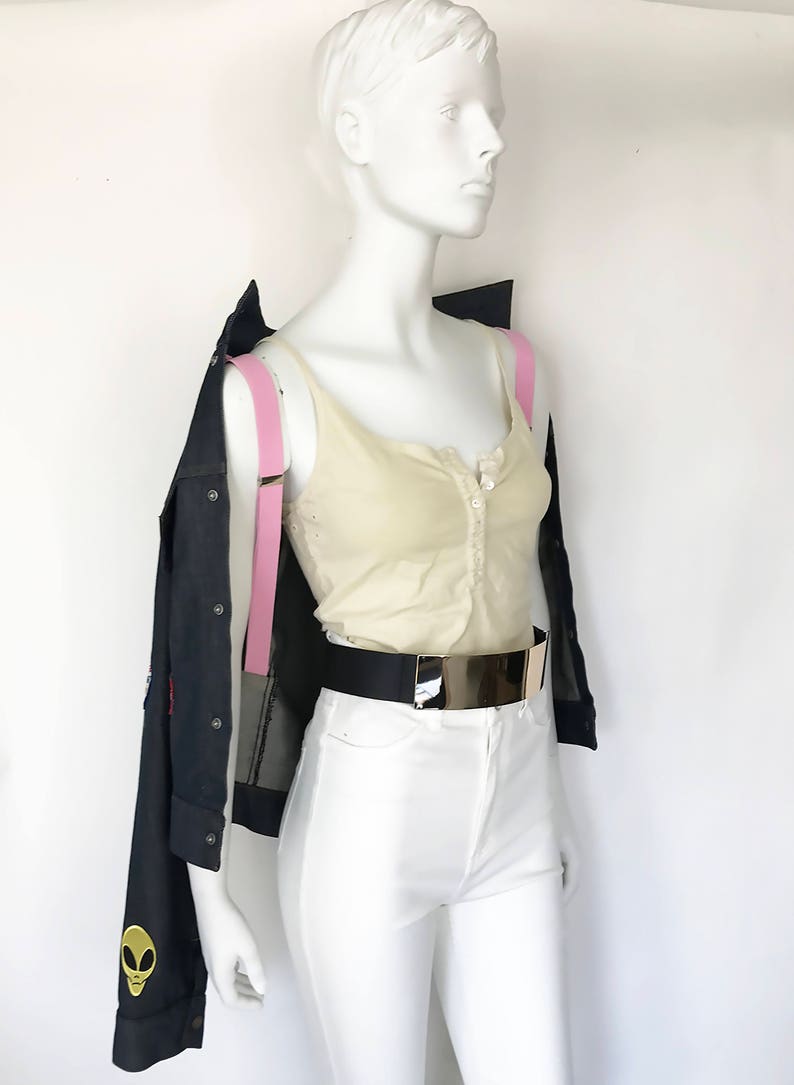 Peut inclure: Un mannequin portant un d&eacute;bardeur blanc, un pantalon blanc, une ceinture noire avec une boucle dor&eacute;e et une veste en jean bleu fonc&eacute; avec des bretelles roses. La veste a un motif extraterrestre sur le c&ocirc;t&eacute; gauche.