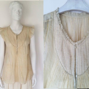 Puede incluir: Una blusa de color blanco roto transparente con cuello de volantes y cierre de botones en la parte delantera. La blusa tiene una textura delicada y brillante y un diseño ligeramente fruncido.