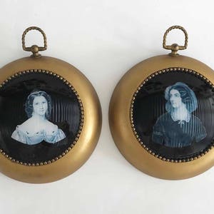 May include: Two antique portrait medallions. Each features a blue and white portrait of a woman set against a black background, framed by a gold-toned metal. The medallions have a decorative loop at the top for hanging.