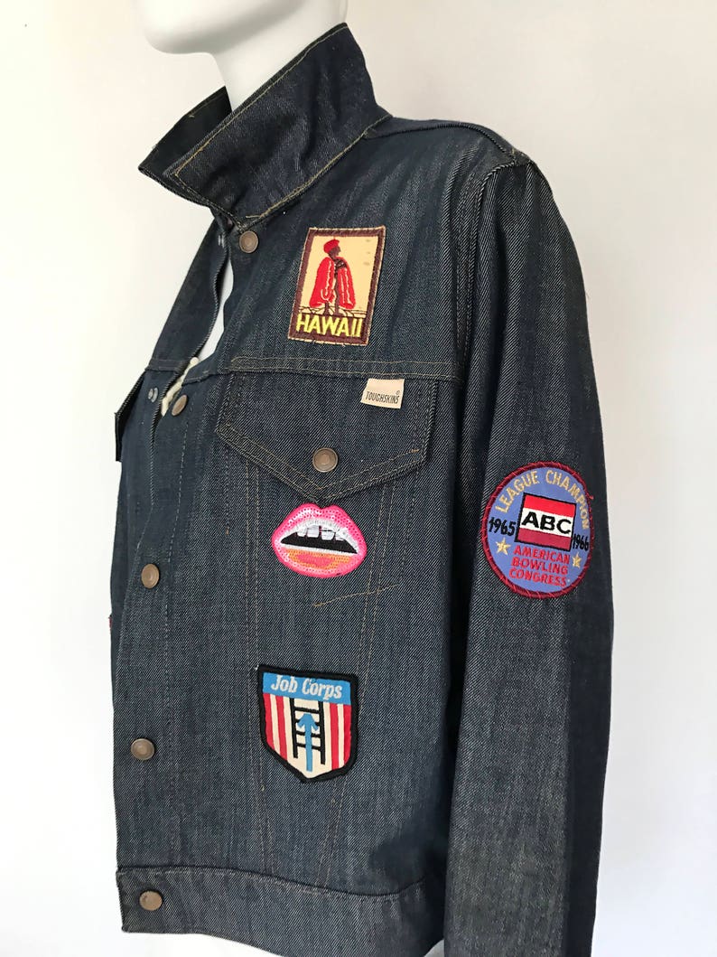 Peut inclure: Une veste en jean bleu fonc&eacute; avec divers patchs, dont un patch "Job Corps" rouge et blanc, un patch bouche rose et blanc, un patch "Hawaii" rouge et blanc et un patch rond avec le texte "1965 ABC 1966 American Bowling Congress League Champion".