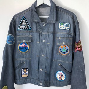 K&ouml;nnte beinhalten: Eine blaue Jeansjacke mit verschiedenen Patches, darunter "LEE TIRES", "CALIFORNIA", "WHITE PASS", "BLUE GOLD", "1962 World's Fair" und ein Schmetterling.