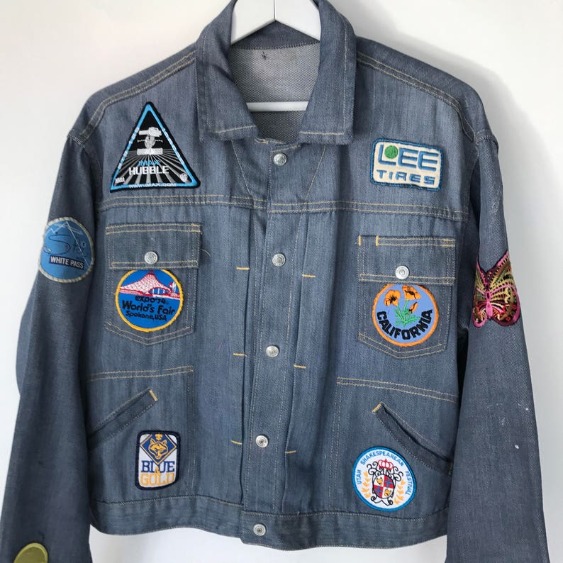 K&ouml;nnte beinhalten: Eine blaue Jeansjacke mit verschiedenen Patches, darunter "LEE TIRES", "CALIFORNIA", "WHITE PASS", "BLUE GOLD", "1962 World's Fair" und ein Schmetterling.