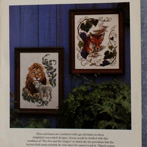 May include: Two framed cross-stitch designs, one depicting a lion and lamb, the other a fox and grapes. The frames are brown, set against a blue background. Text at the top reads "Fabled Pair".
