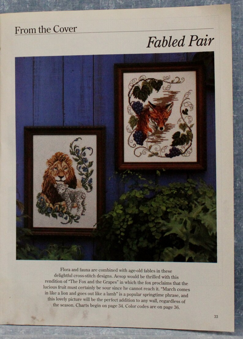 May include: Two framed cross-stitch designs, one depicting a lion and lamb, the other a fox and grapes. The frames are brown, set against a blue background. Text at the top reads "Fabled Pair".