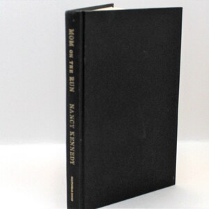 Puede incluir: Un libro de tapa dura negro con el t&iacute;tulo "MOM ON THE RUN" y el autor "NANCY KENNEDY" impreso en dorado en el lomo.