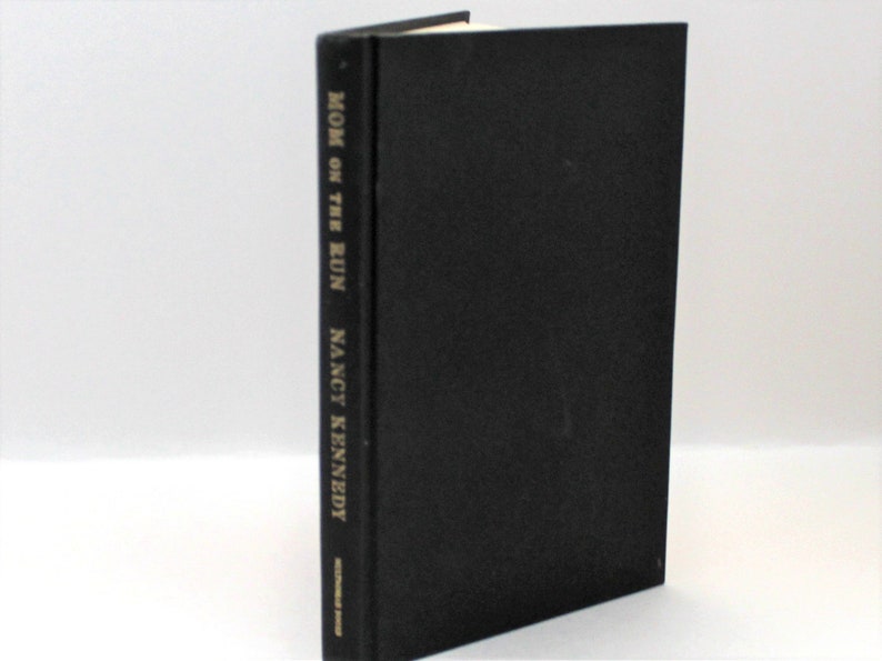 Puede incluir: Un libro de tapa dura negro con el t&iacute;tulo "MOM ON THE RUN" y el autor "NANCY KENNEDY" impreso en dorado en el lomo.