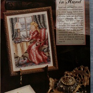 May include: Cross-stitch picture in a gold frame showing a woman in a red dress writing at a desk. The image includes an ornate inkwell and books. The text on the image reads "With Pen in Hand". The artwork is displayed on a dark surface.