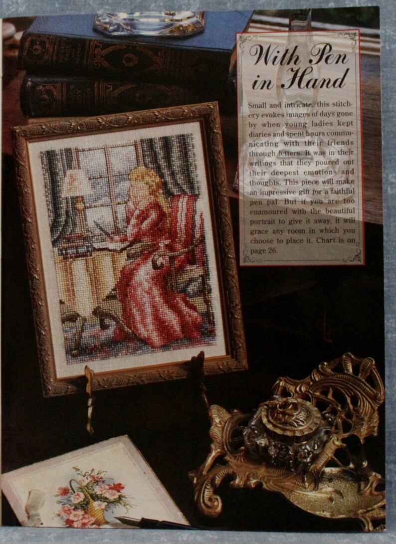 May include: Cross-stitch picture in a gold frame showing a woman in a red dress writing at a desk. The image includes an ornate inkwell and books. The text on the image reads "With Pen in Hand". The artwork is displayed on a dark surface.