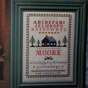 May include: A framed cross-stitch sampler with a house, trees, and the alphabet. The sampler is titled "MOORE" and includes the text "wrought by Anne Moore in 1987".