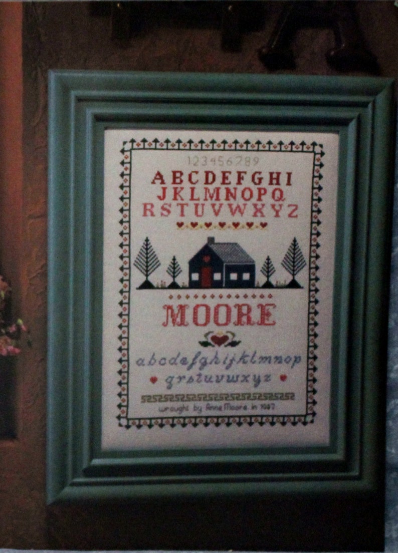 May include: A framed cross-stitch sampler with a house, trees, and the alphabet. The sampler is titled "MOORE" and includes the text "wrought by Anne Moore in 1987".