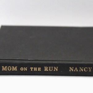 Puede incluir: Un libro negro con letras doradas en el lomo que dice "MOM ON THE RUN" de "NANCY KENNEDY".