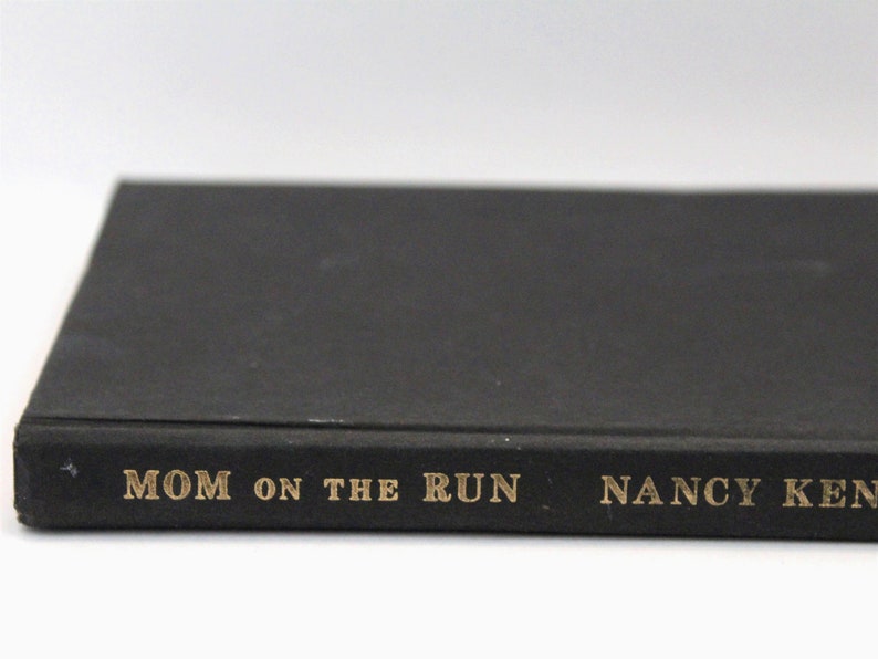 Puede incluir: Un libro negro con letras doradas en el lomo que dice "MOM ON THE RUN" de "NANCY KENNEDY".