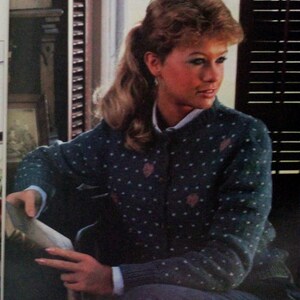 May include: A woman wearing a blue and white patterned cardigan with a white button-up shirt underneath. She is sitting in front of a window with blinds.