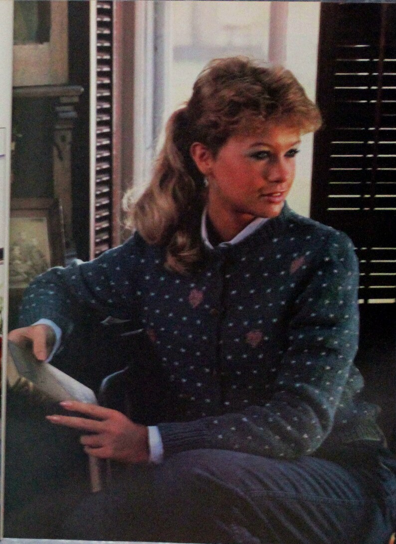 May include: A woman wearing a blue and white patterned cardigan with a white button-up shirt underneath. She is sitting in front of a window with blinds.