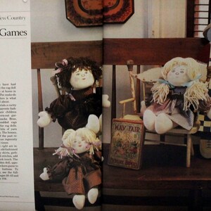 May include: Three rag dolls, one sitting on a wooden chair, one sitting on a wooden bench, and one standing. The dolls are dressed in brown and white checkered fabric and have yarn hair. The doll standing is wearing a blue and white checkered apron. The dolls are surrounded by wooden toys and a book titled "May Fair."
