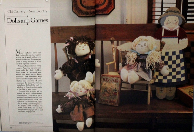 May include: Three rag dolls, one sitting on a wooden chair, one sitting on a wooden bench, and one standing. The dolls are dressed in brown and white checkered fabric and have yarn hair. The doll standing is wearing a blue and white checkered apron. The dolls are surrounded by wooden toys and a book titled "May Fair."